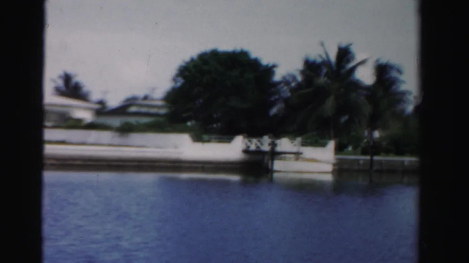 A 1955 Dade County canal-side view with tropical landscaping, a white shoreline wall, palms, and blue water in the foreground.