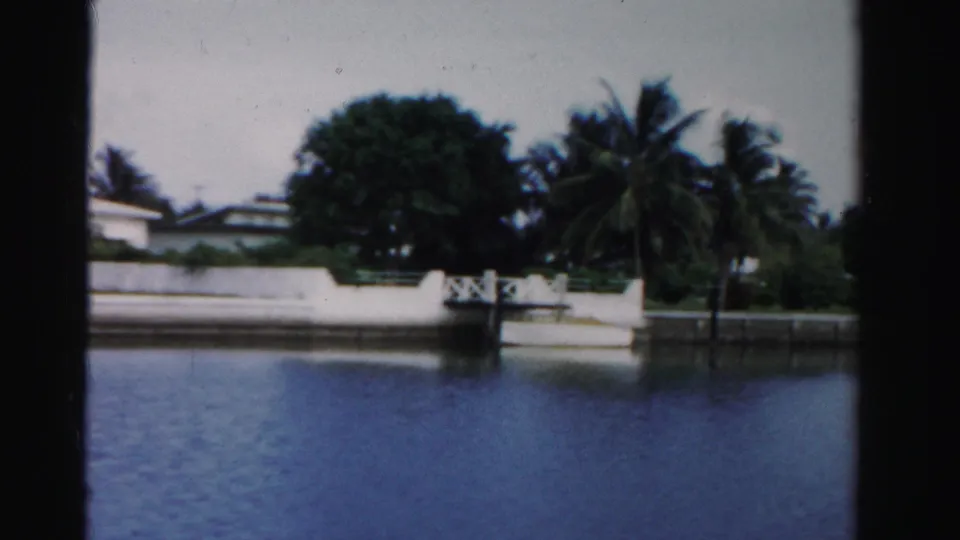 1955 Dade County canal view with blue water, white retaining walls, palm trees, and low waterfront homes.