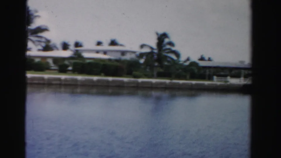 Dade County waterfront houses and palms appear across the canal, with soft 8mm color and a concrete seawall along the shore.