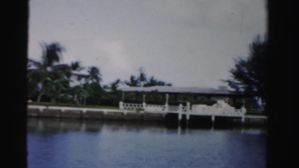 A white dock railing appears along the Dade County canal, with low homes, palms, and dense greenery in the background.