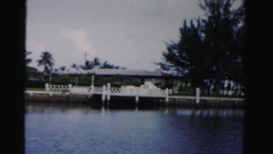 The final Dade County frame shows a white dock and railing, calm canal water, palms, and a low waterfront house partly screened by trees.