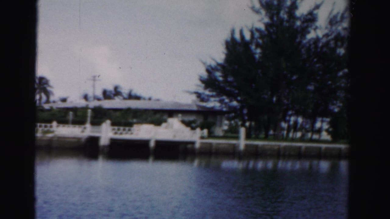 White waterfront house with dock, palms, and calm water seen through dark film-frame edges.