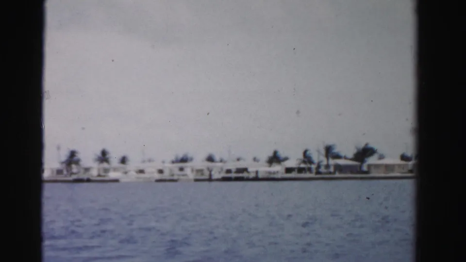 Distant 1955 Dade County waterfront homes and palms line the shore across blue water in faded 8mm color.