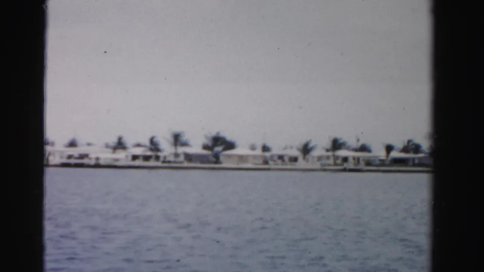 1955 Dade County 8mm frame showing open water with low white homes and palm trees on the far shore.