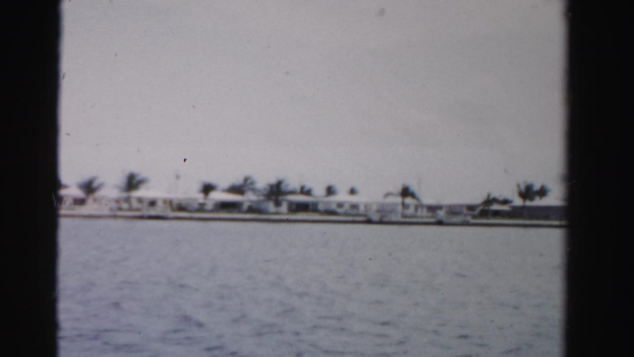 1955 Dade County 8mm frame showing low waterfront buildings and palms across a broad waterway under a gray sky.
