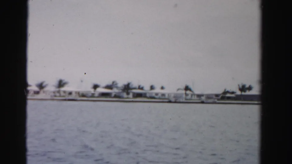 1955 Dade County 8mm frame showing low waterfront buildings and palms across a broad waterway under a gray sky.