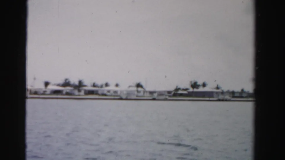 Dade County waterfront view on 8mm film, with light buildings, palms, and a seawall visible across the water.