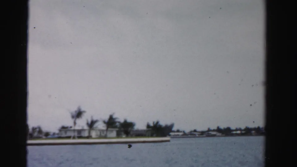 Soft 1955 8mm view of a Dade County shoreline with palms, low buildings, and water in the foreground.