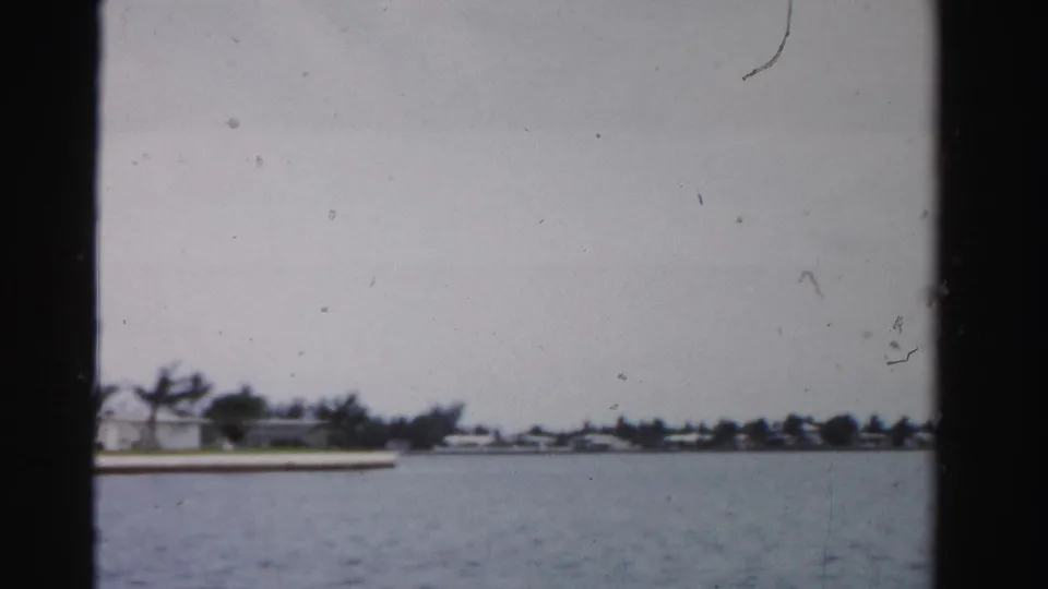 1955 Dade County, Florida waterway viewed from a boat, with distant shoreline houses, trees, overcast sky, and visible 8mm film borders.