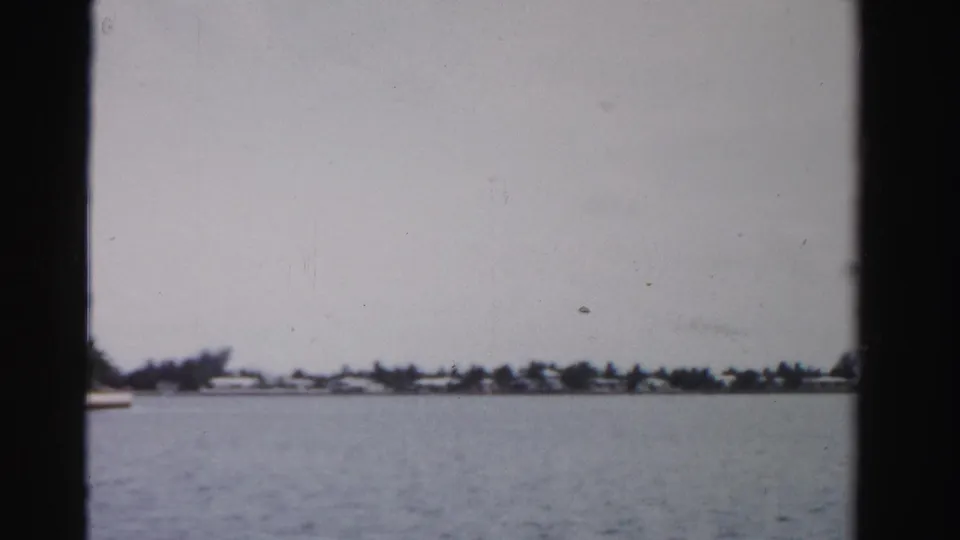 Boat-view frame of a Dade County waterway in 1955, showing open water, low houses, palms, gray sky, and soft 8mm grain.