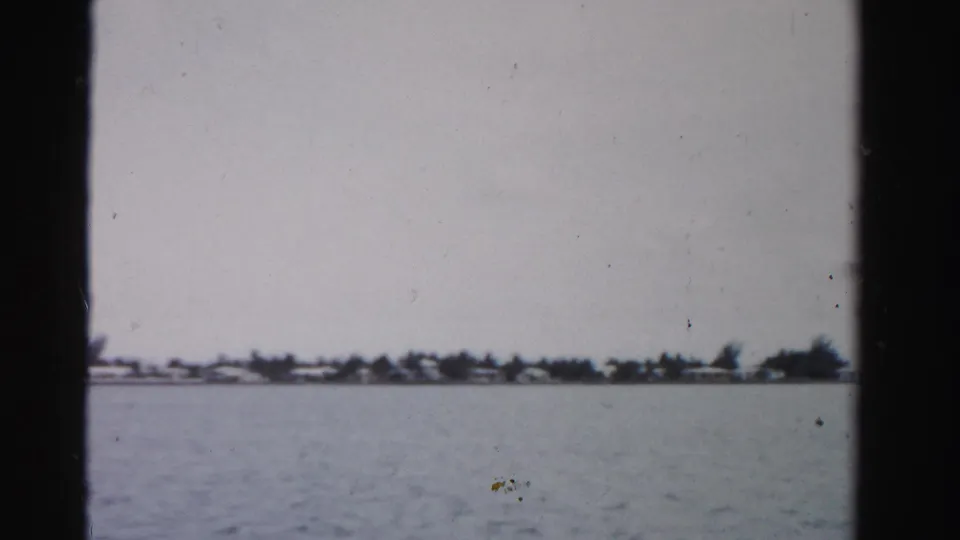 1955 home-movie view across a Florida waterway toward a low residential shoreline with trees, pale sky, and 8mm film wear.
