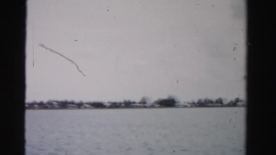 Final 1955 Dade County waterway frame with calm water, a thin distant shore, blurred coastal trees, visible scratches, and 8mm borders.