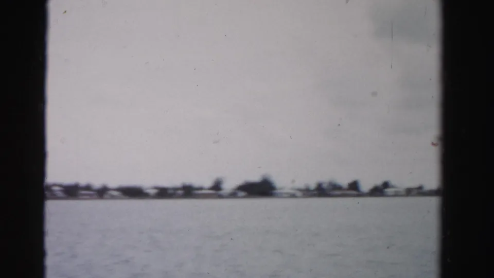 1955 Dade County 8mm frame showing open water and a low distant shoreline with homes and trees under an overcast sky.