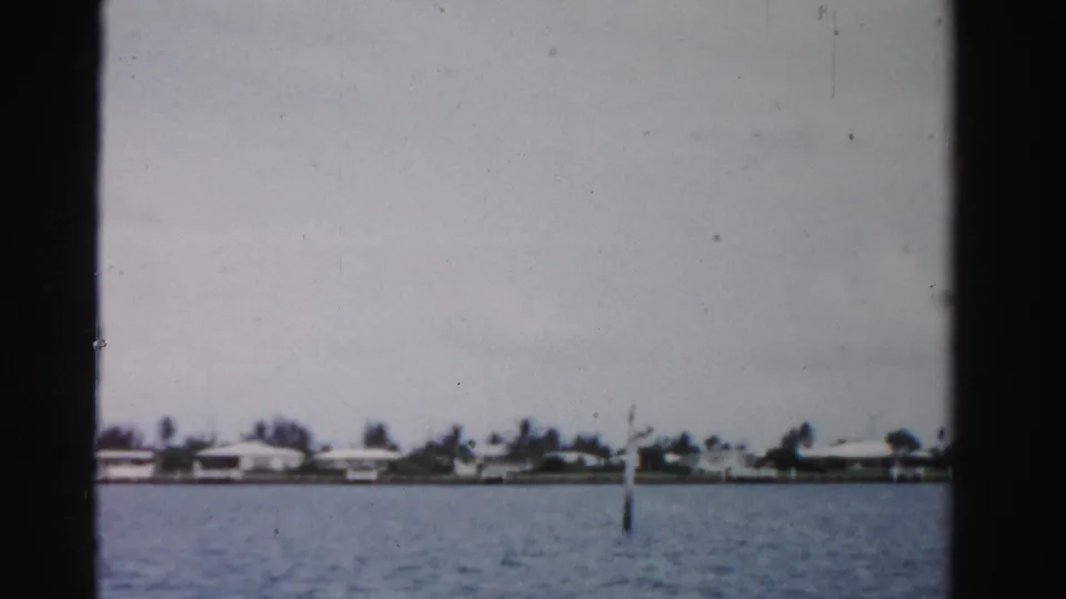 1955 Dade County 8mm frame of rippled water, a distant residential shoreline, trees, and a small marker post near the center.