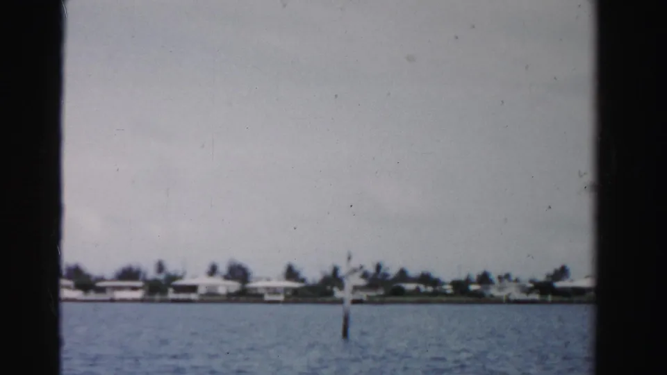 1955 Dade County 8mm frame showing a calm waterway, distant palm-lined shoreline, low waterfront buildings, and a marker pole.