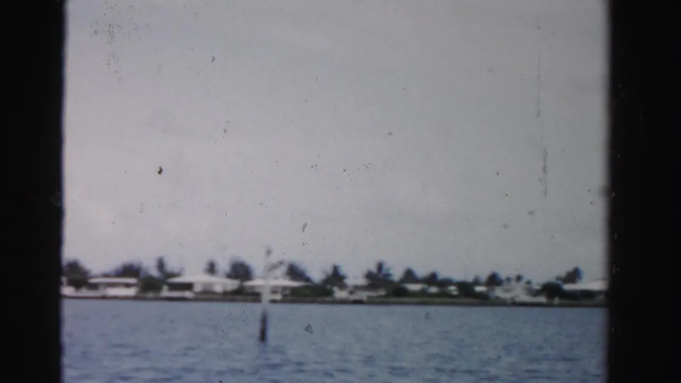 1955 Dade County boat-view frame with calm water, a distant residential-looking shoreline, palm trees, and a single water marker.