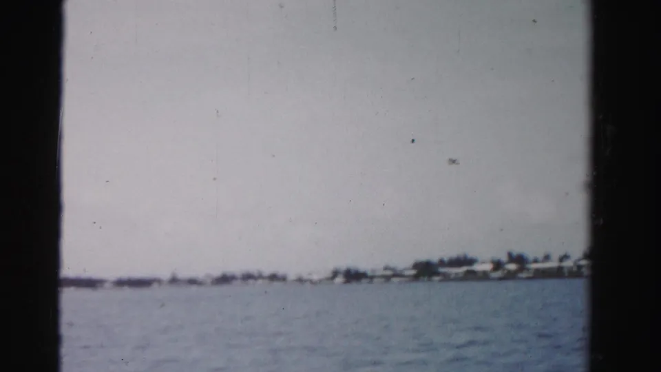 1955 Dade County 8mm frame showing open water from a boat and a low, distant shoreline with small waterfront structures.
