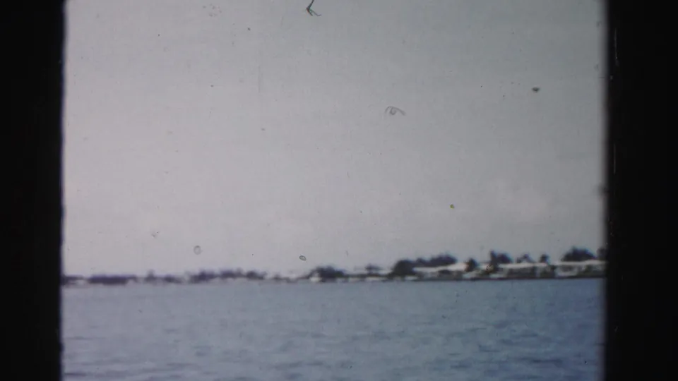 1955 Dade County boat-trip frame showing calm water, a far palm-lined shore, and low waterfront buildings beneath a pale sky.