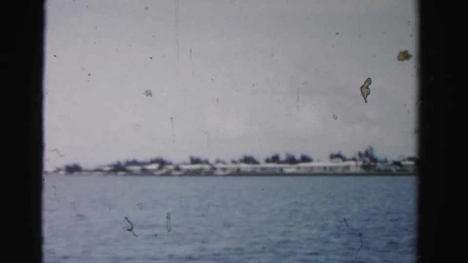 Mid-century Dade County shoreline seen across open water, with low houses and a soft, scratched 8mm film surface.