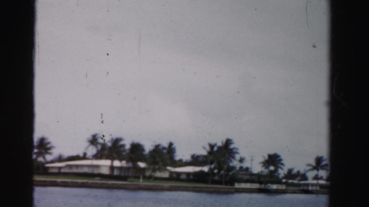 1955 Dade County home-movie frame showing white waterfront residences, palms, seawall, and calm water viewed from across a canal.