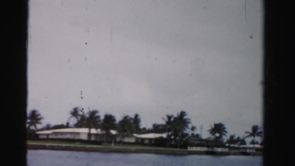 1955 Dade County home-movie frame showing white waterfront residences, palms, seawall, and calm water viewed from across a canal.