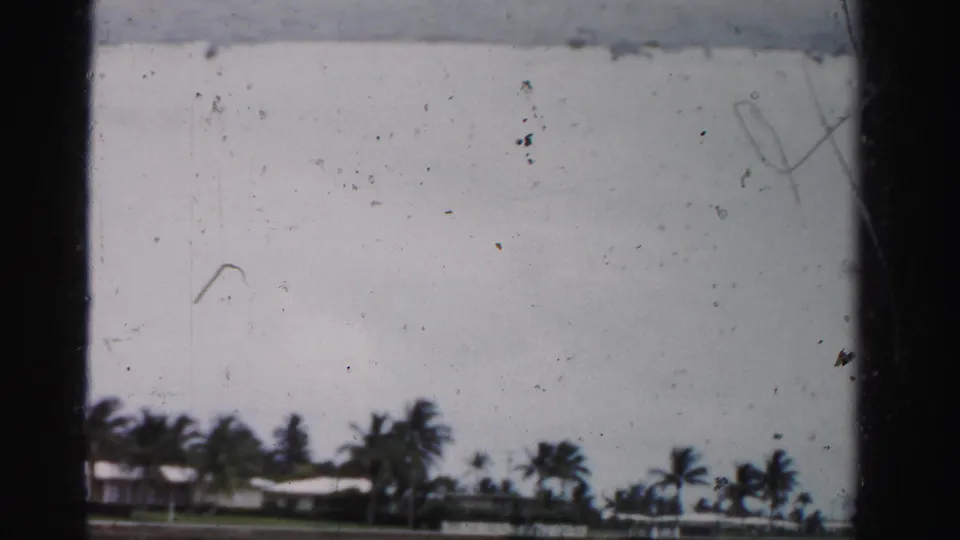 Soft 1955 home-movie frame of low waterfront homes, palms, seawall, and pale sky in Dade County, Florida.