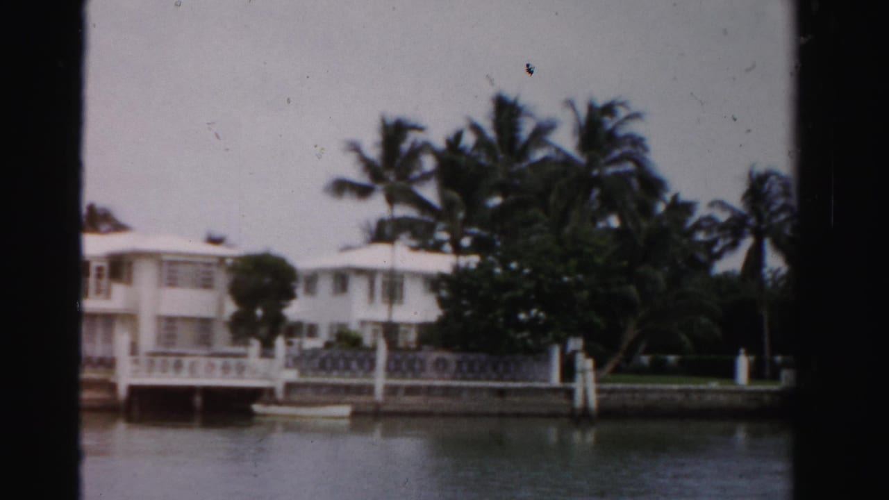 1955 Dade County 8mm frame showing a white canal-front house, palm trees, low railing, dock posts, and calm water.