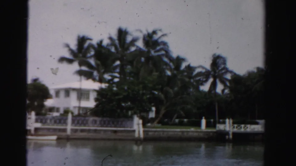 1955 Dade County waterfront view with white houses, palm trees, dock posts, railing, and still canal water.