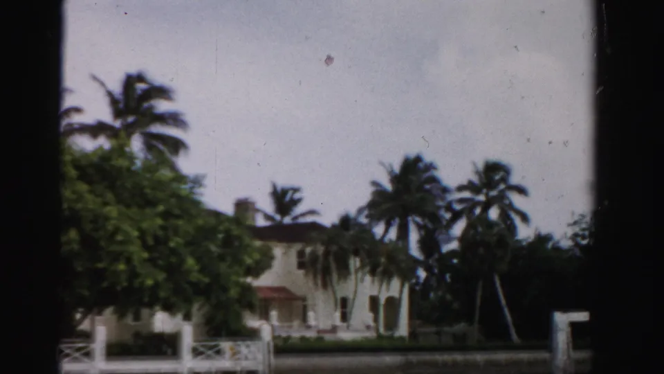 1955 Dade County 8mm frame showing a waterfront residence with palms, leafy tree cover, railing, dock posts, and canal water.