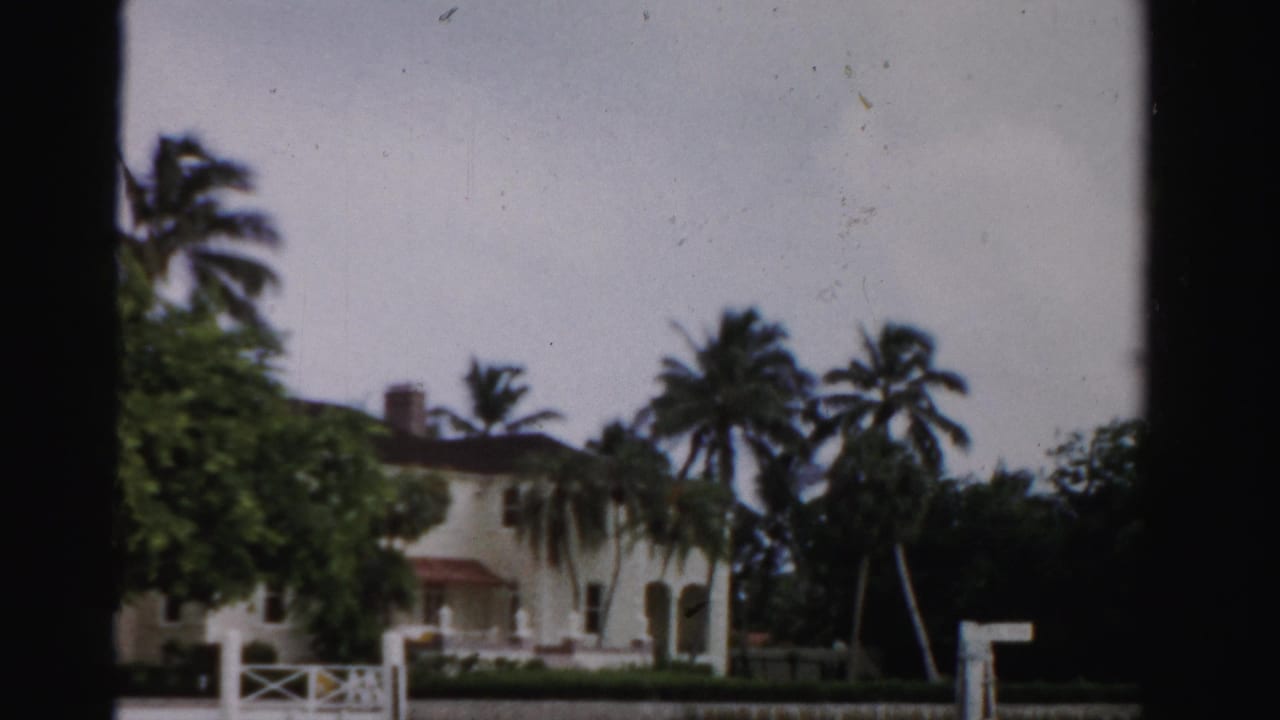 1955 Dade County, Florida home-movie view of a white stucco residence with palms, lawn, and low fencing behind dark foreground framing.