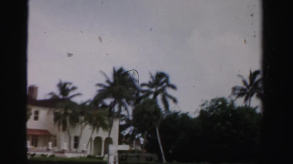 The home-movie frame shows the left side of a white Florida residence, palm trees, shrubs, and a low fence under a pale sky.