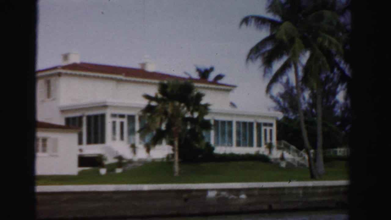 1955 Dade County waterfront view of a white stucco house with red tile roof, green lawn, palm trees, seawall, and water.