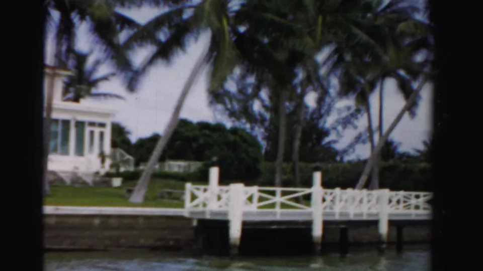 End of the waterfront pass showing a white dock, leaning palm trees, seawall, water, lawn, and the edge of the Dade County house.