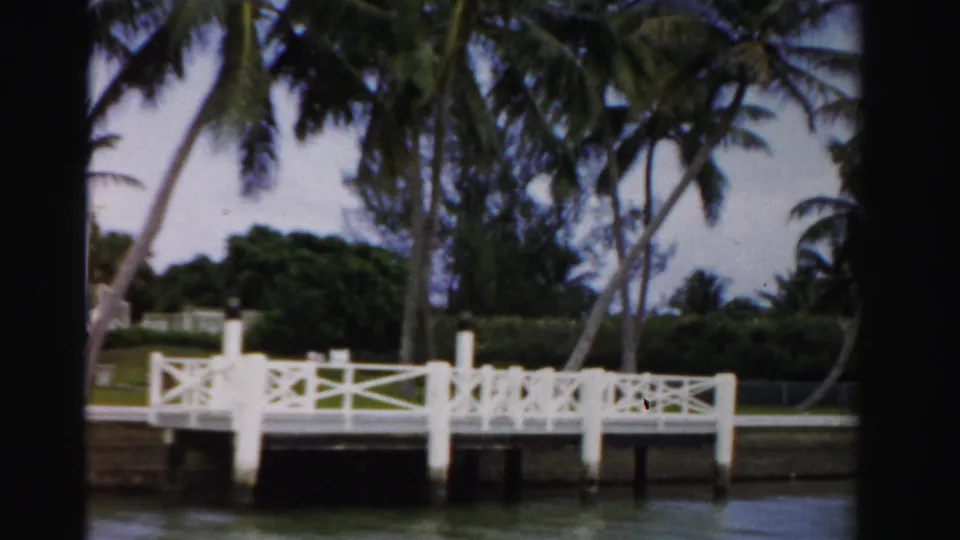 1955 Dade County 8mm view of a white canal-side landing, palm trees, hedges, and lawn seen from across the water.