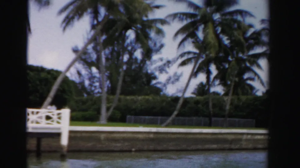 The landing slips partly out of frame as the 8mm camera continues past palms, a lawn, hedges, and canal water in Dade County.
