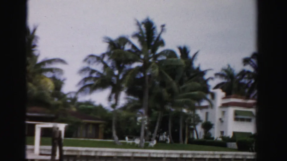 1955 Dade County waterfront estate scene with palms, a white house, dock railing, lawn, and seawall.