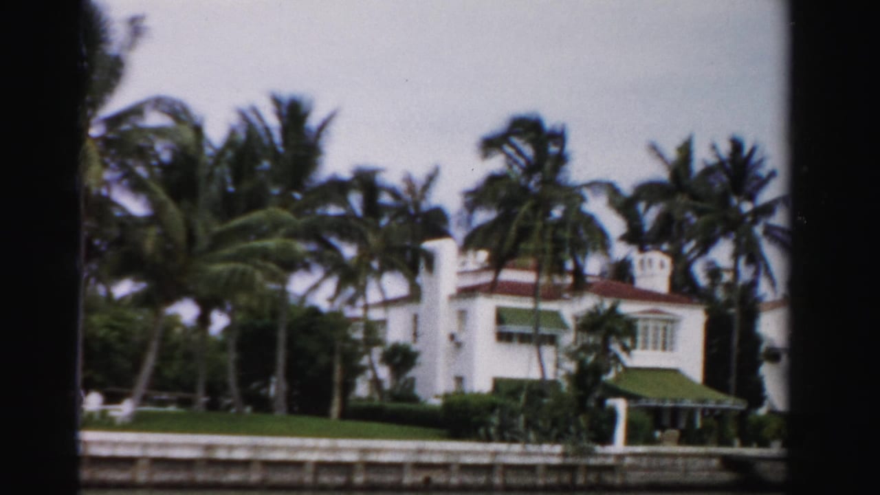 1955 Dade County home-movie frame showing a white Mediterranean-style house, palms, green lawn, and canal frontage.
