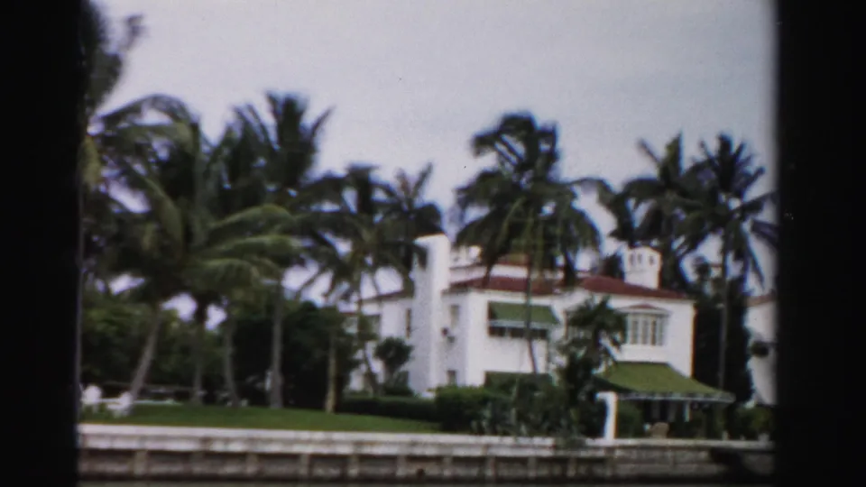 1955 Dade County home-movie frame showing a white Mediterranean-style house, palms, green lawn, and canal frontage.