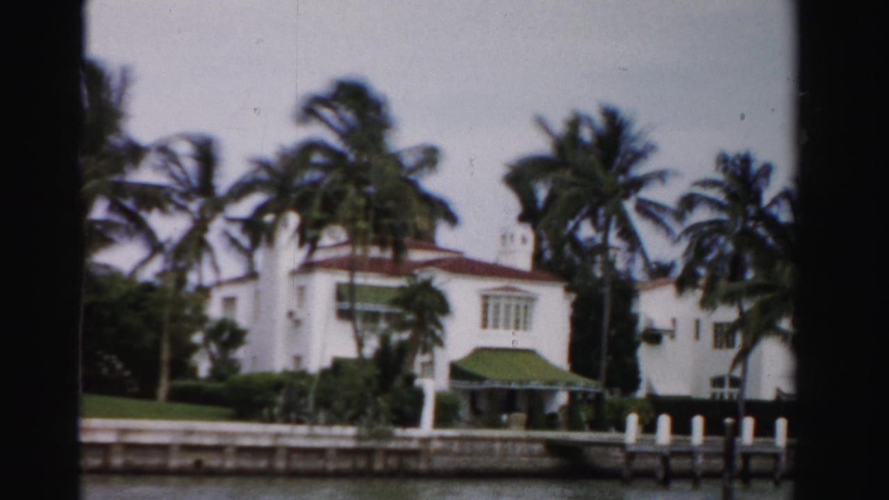 1955 Dade County frame of a white waterfront residence with palms, awning, dock posts, and a canal in front.