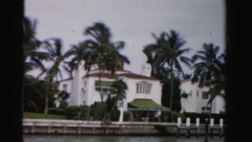 1955 Dade County frame of a white waterfront residence with palms, awning, dock posts, and a canal in front.