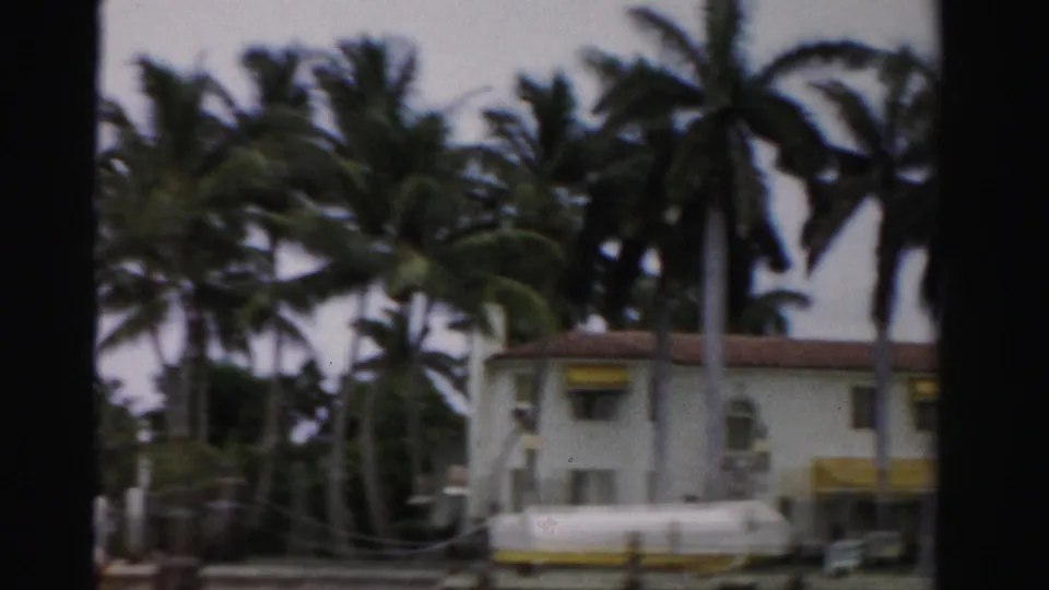 1955 Dade County home-movie frame showing a low white waterfront building with yellow awnings behind tall palms.