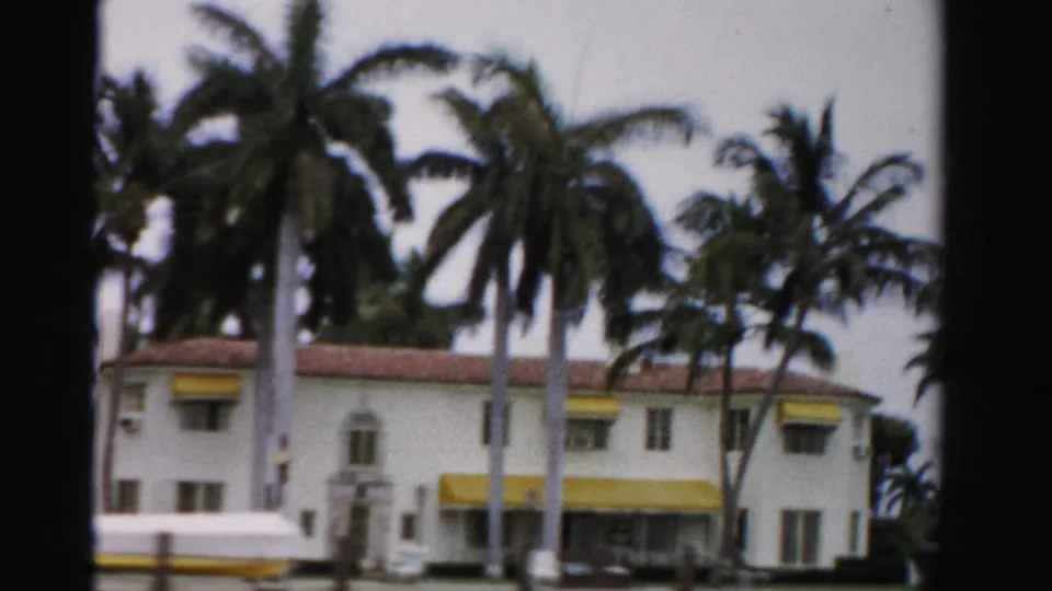 1955 Dade County frame showing a white canal-side building with yellow awnings, red tile roof, and clustered palms.