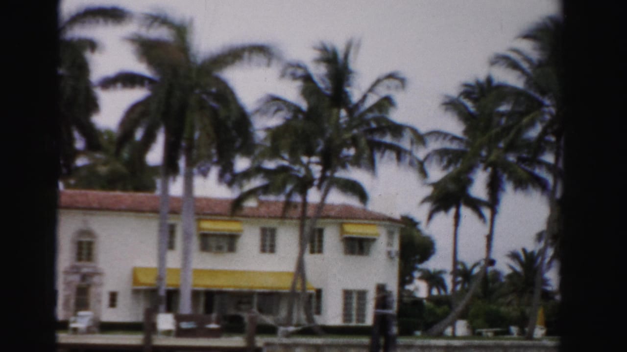 1955 Dade County home-movie view of a white stucco house with yellow awnings, palms, a hedge, and cloudy sky across water.