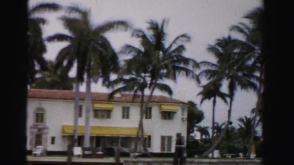 1955 Dade County home-movie view of a white stucco house with yellow awnings, palms, a hedge, and cloudy sky across water.