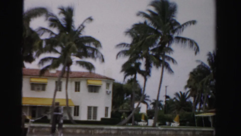 Mid-1950s Dade County footage shows a white house with yellow awnings, palms, hedge line, and a low waterfront wall.