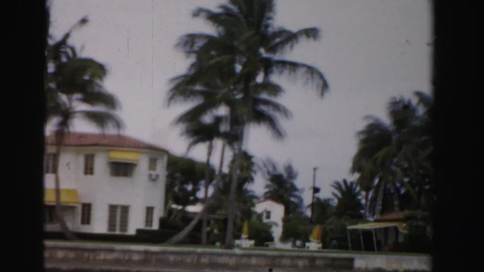 1955 Dade County view from a moving camera, showing the side of a white house, palms, hedge, waterfront wall, and nearby shelter.