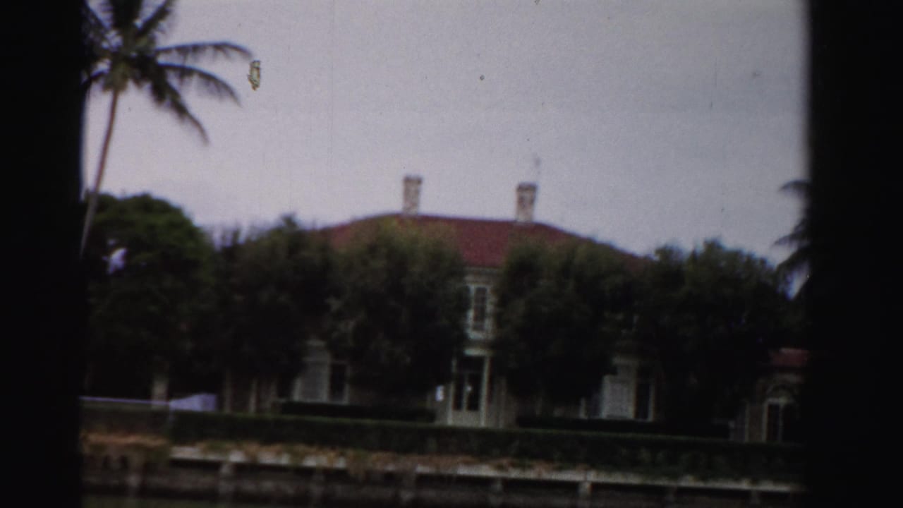 1955 Dade County 8mm frame showing a canal-side house with red tile roof, palms, hedges, and dark projector-frame edges.