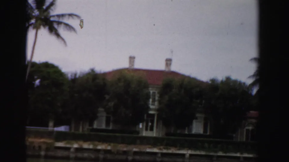 1955 Dade County 8mm frame showing a canal-side house with red tile roof, palms, hedges, and dark projector-frame edges.