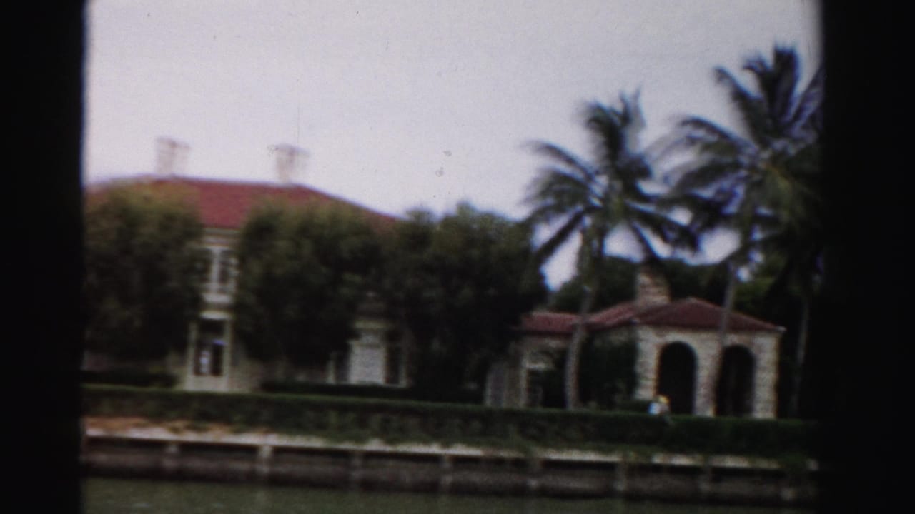 Large waterfront home, palms, hedges, and arched porch beside a canal.