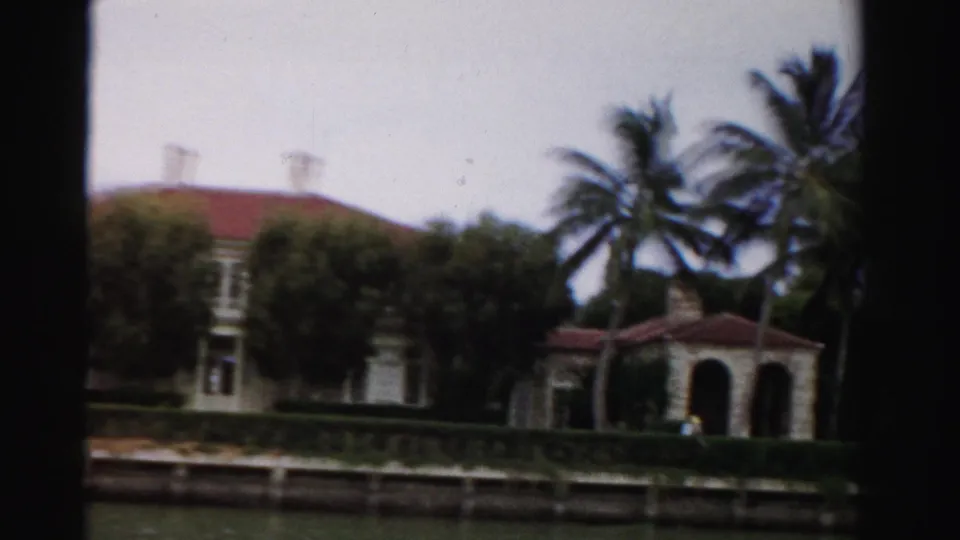 1955 Dade County frame with a large stucco waterfront home, arched porch, red roof, palms, and manicured hedge by the canal.